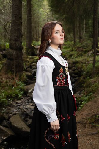 A traditional Norwegian folk costume with red embroidery is showcased in a serene forest setting along a woodland path.