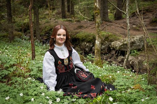A person in traditional Norwegian folk dress sits among white wildflowers in a lush forest setting.
