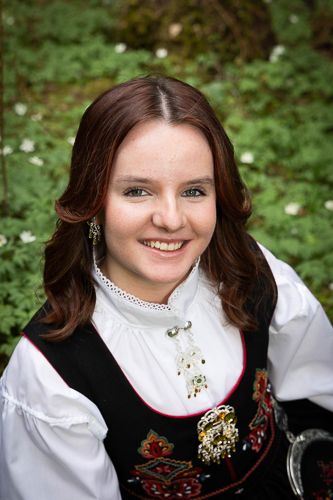 A smiling person wearing traditional Norwegian folk dress with a white blouse and embroidered black vest in an outdoor setting.