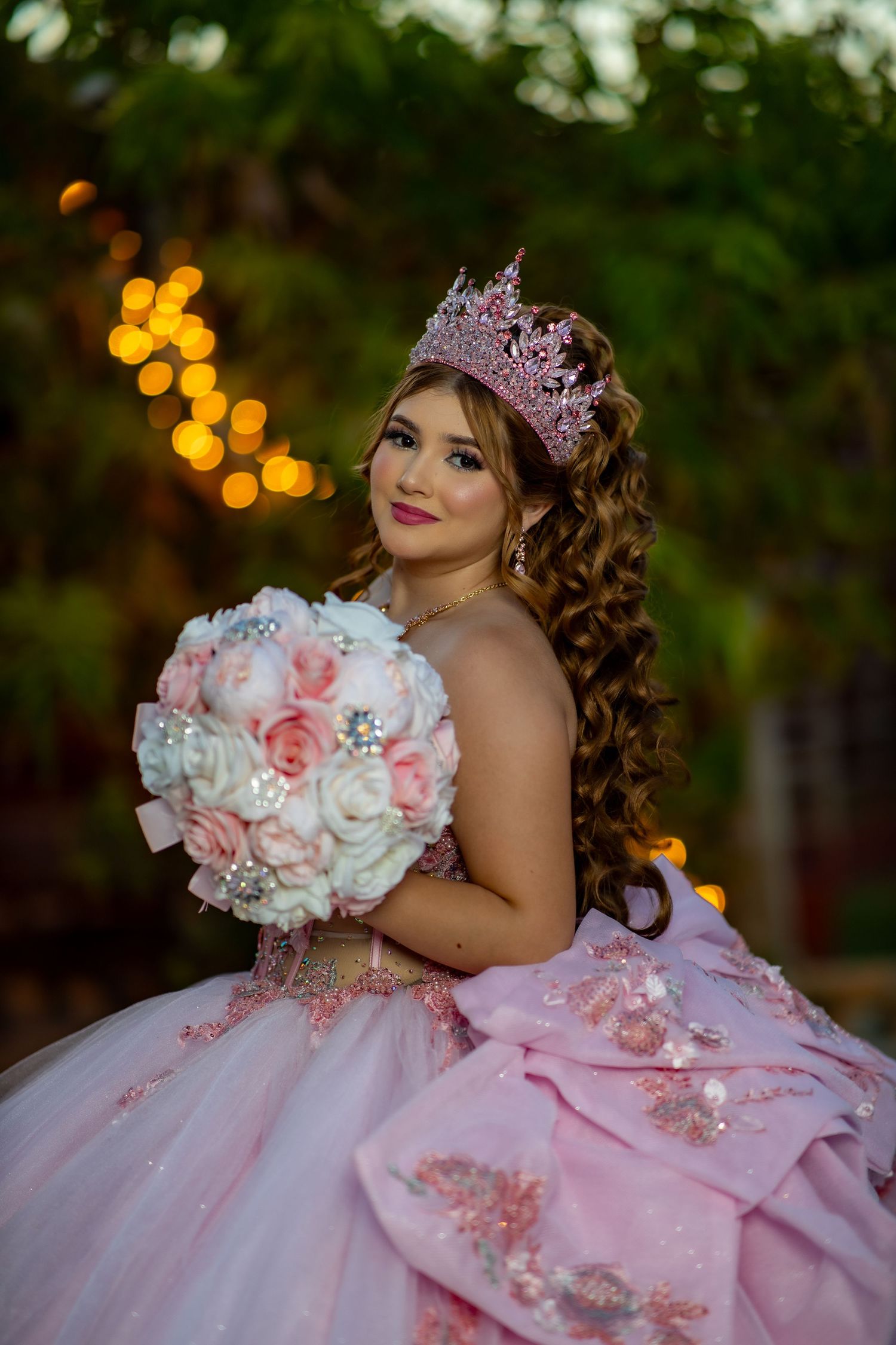 Quinceañera girl in pink gown wearing a crystal crown holds a colorful bouquet while lying on grass.