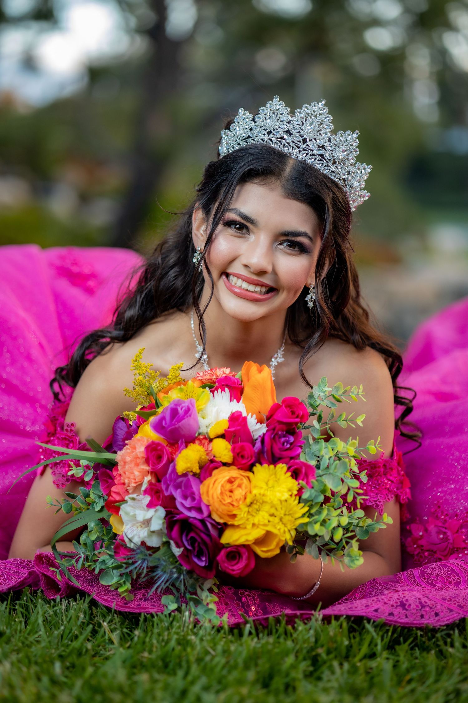 Quinceañera girl in pink gown wearing a crystal crown holds a colorful bouquet while lying on grass.