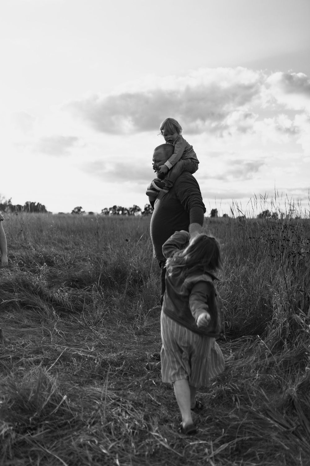 A silhouetted figure walks with a child through a field at sunset against a cloudy sky.