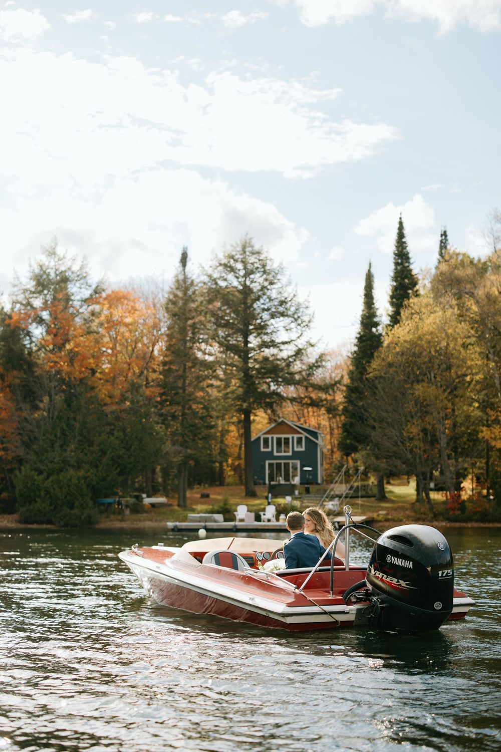 A red motorboat cruises past a lakeside cabin surrounded by autumn trees with vibrant fall foliage.