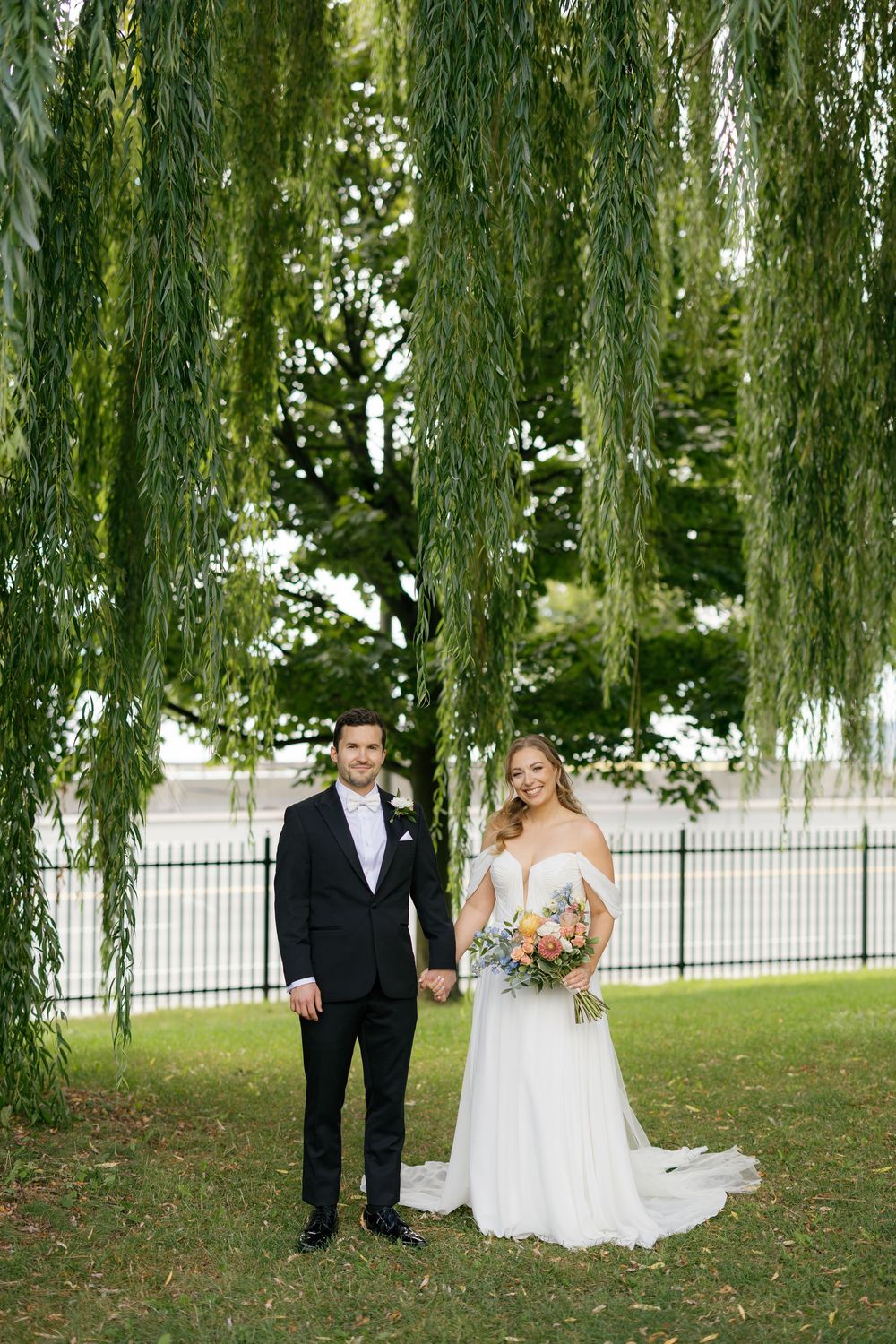 A romantic wedding couple poses under a graceful weeping willow tree in a lush garden setting.
