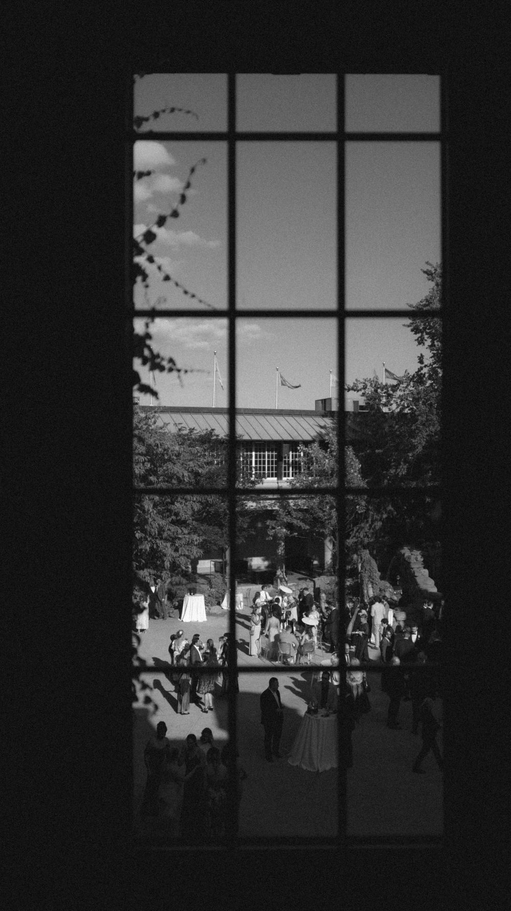 Black and white view through window panes showing silhouettes of people gathering in a courtyard or outdoor space.
