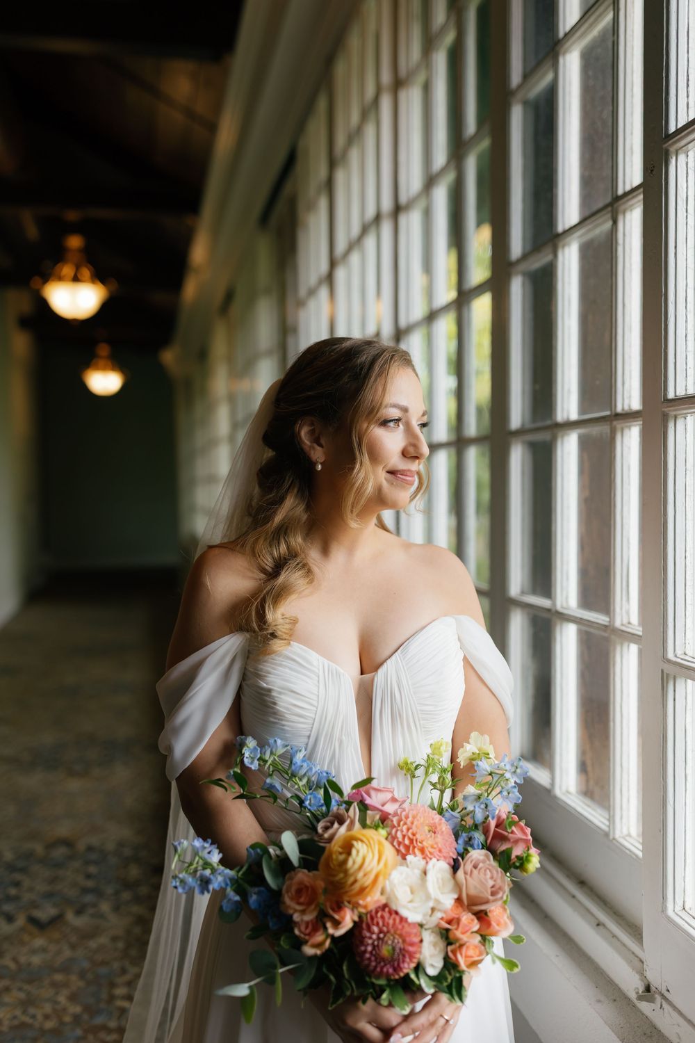 Bride in white dress holds colorful bouquet while gazing through sunlit windows in elegant venue.