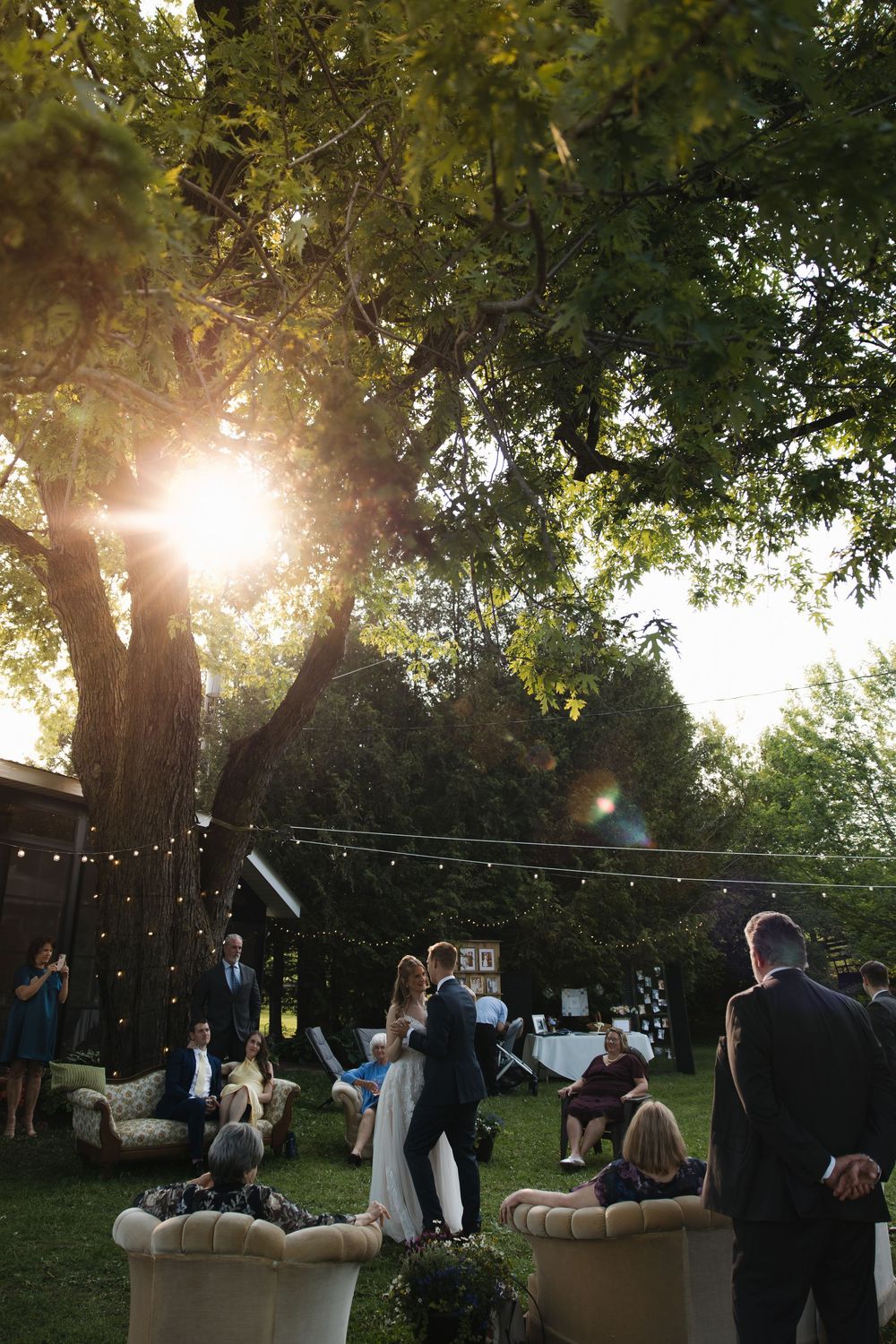 Outdoor wedding ceremony at sunset with guests gathered under large trees they are doing their first dance