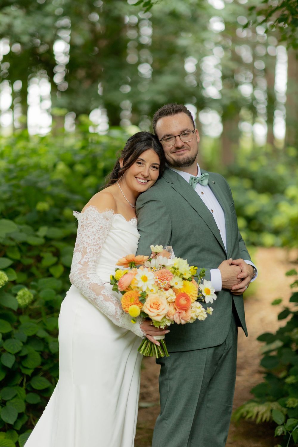 A couple in wedding attire poses together in a lush green forest setting with a vibrant orange and peach floral bouquet.