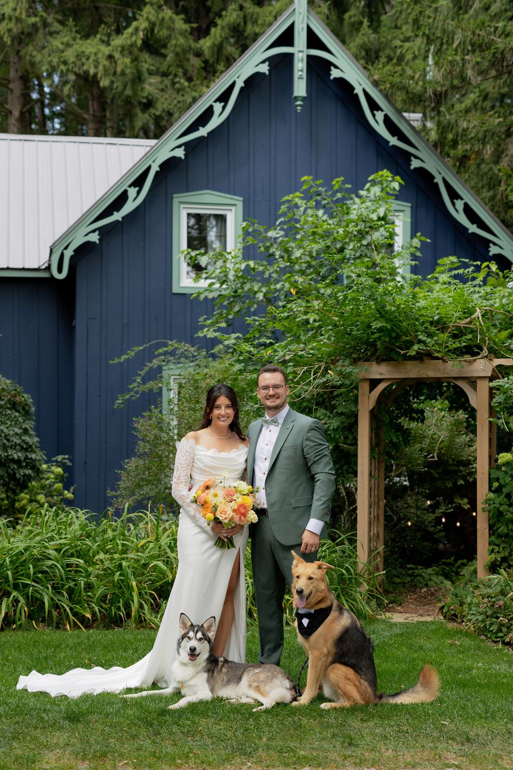 A couple poses with their two dogs in front of a blue barn during a rustic outdoor wedding celebration.