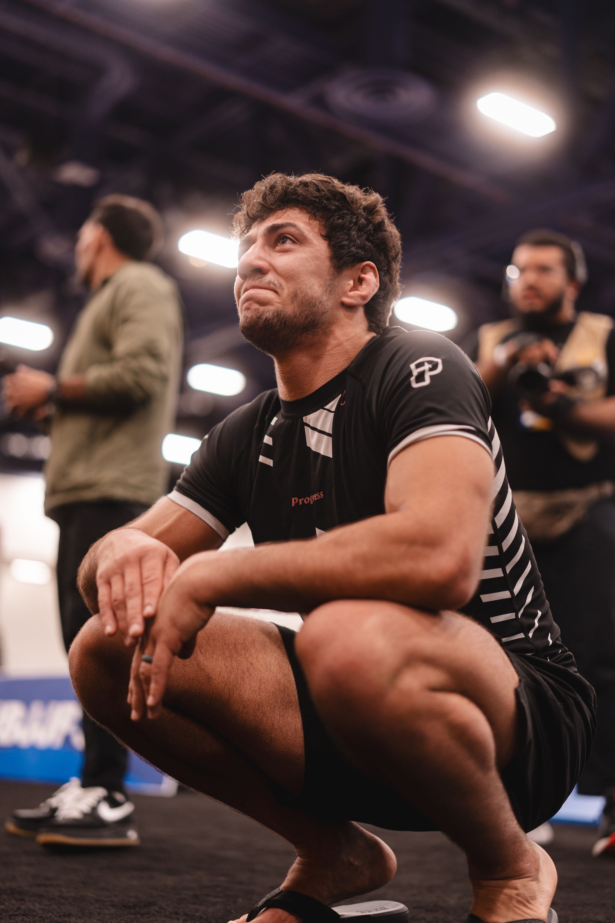 Male BJJ athlete in black rash guard crouching on competition floor looking upward with intense expression.