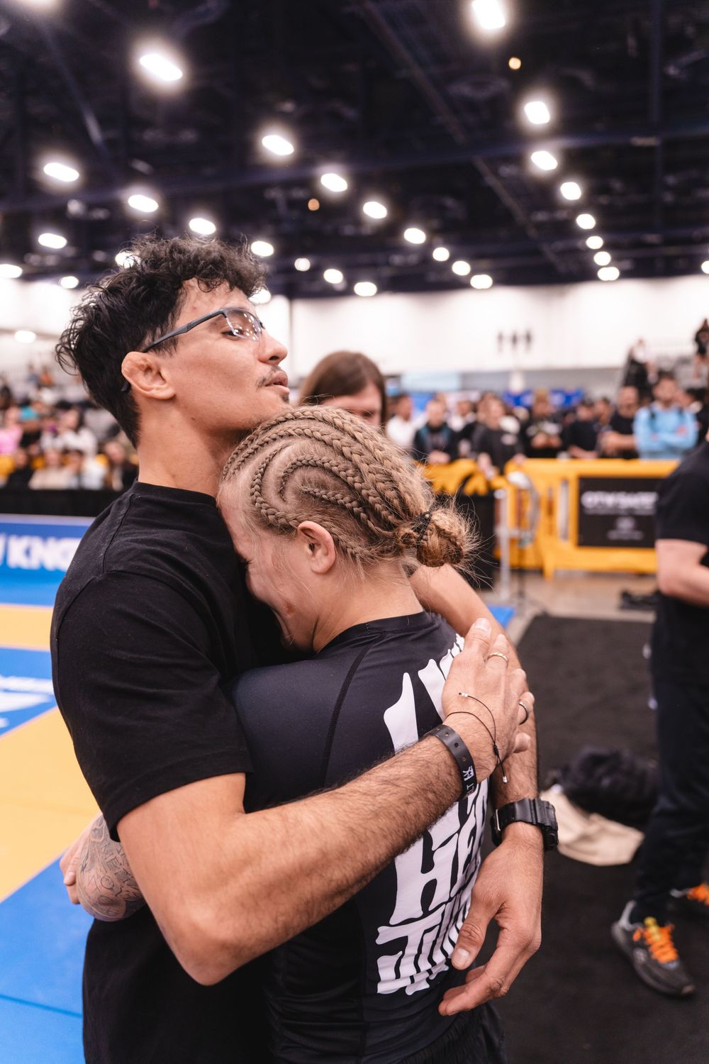 Two athletes in black shirts share an emotional hug at an indoor sports competition with blue mats visible.