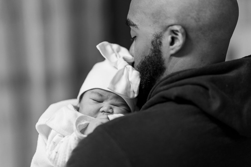 A tender moment shows a baby in white clothing sleeping peacefully against someone's shoulder.