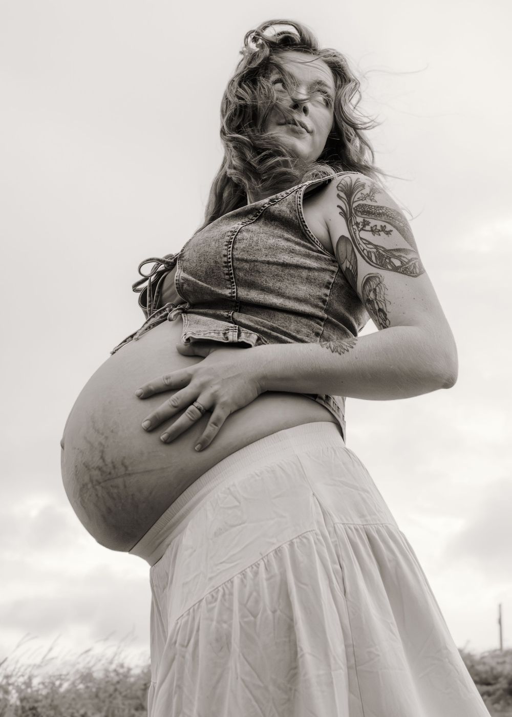 Black and white photo of a pregnant woman with tattoos wearing a denim top and white skirt outdoors.