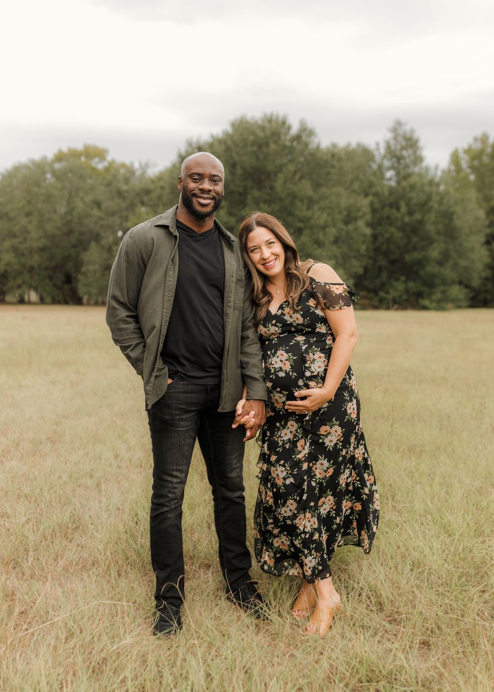 A couple stands together in a grassy field on an overcast day, with trees in the background.