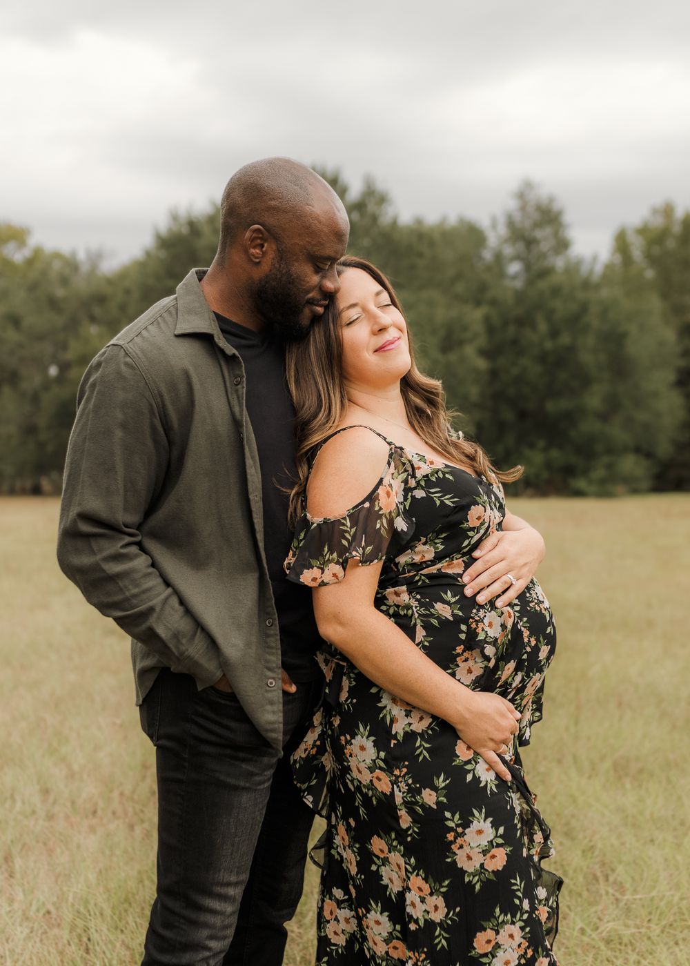 A couple in a field shares an intimate embrace, with her wearing a floral dress and him in dark clothing.