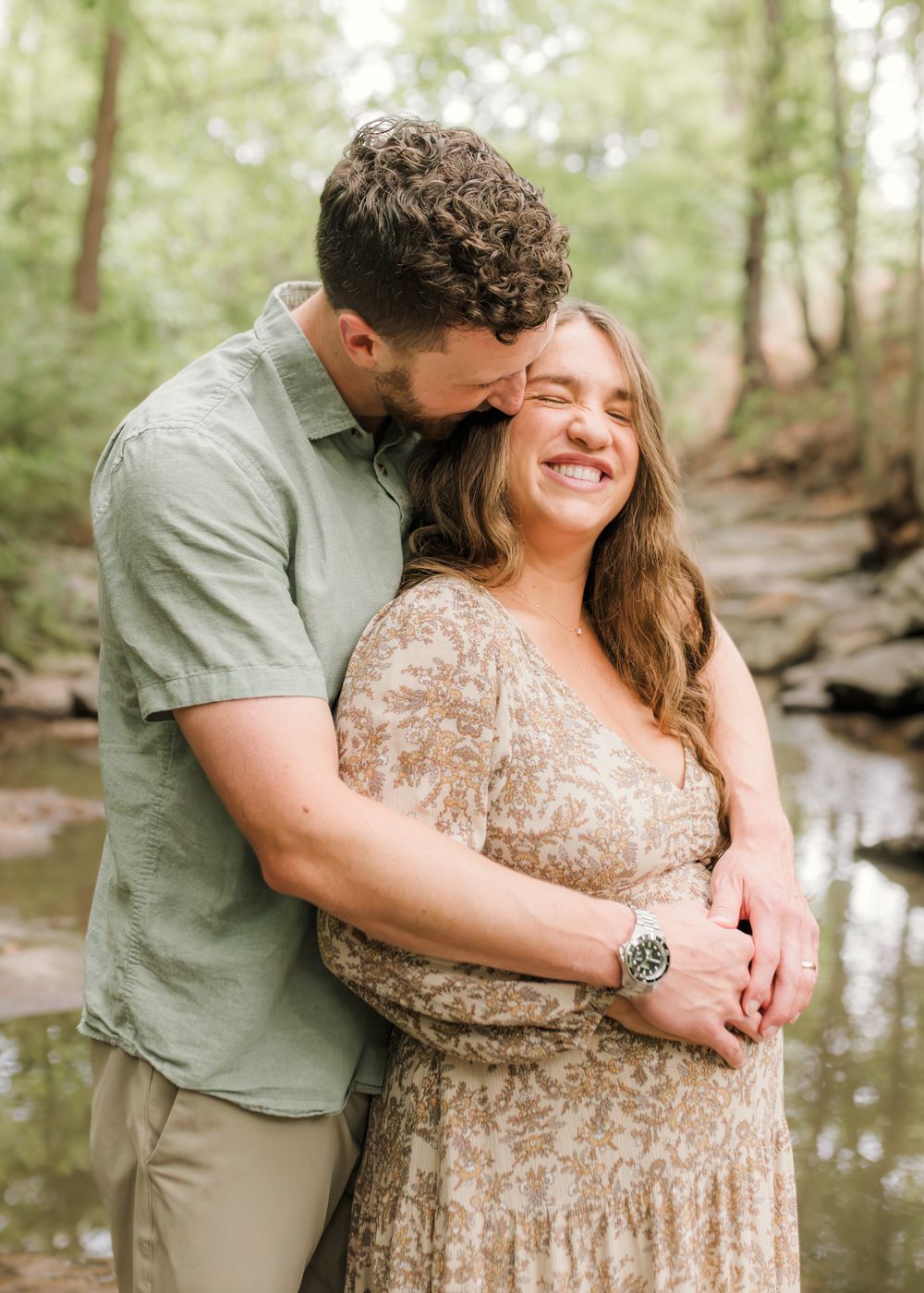 Couple embracing in a forest creek setting, woman smiling joyfully as man kisses her head, engagement photo.