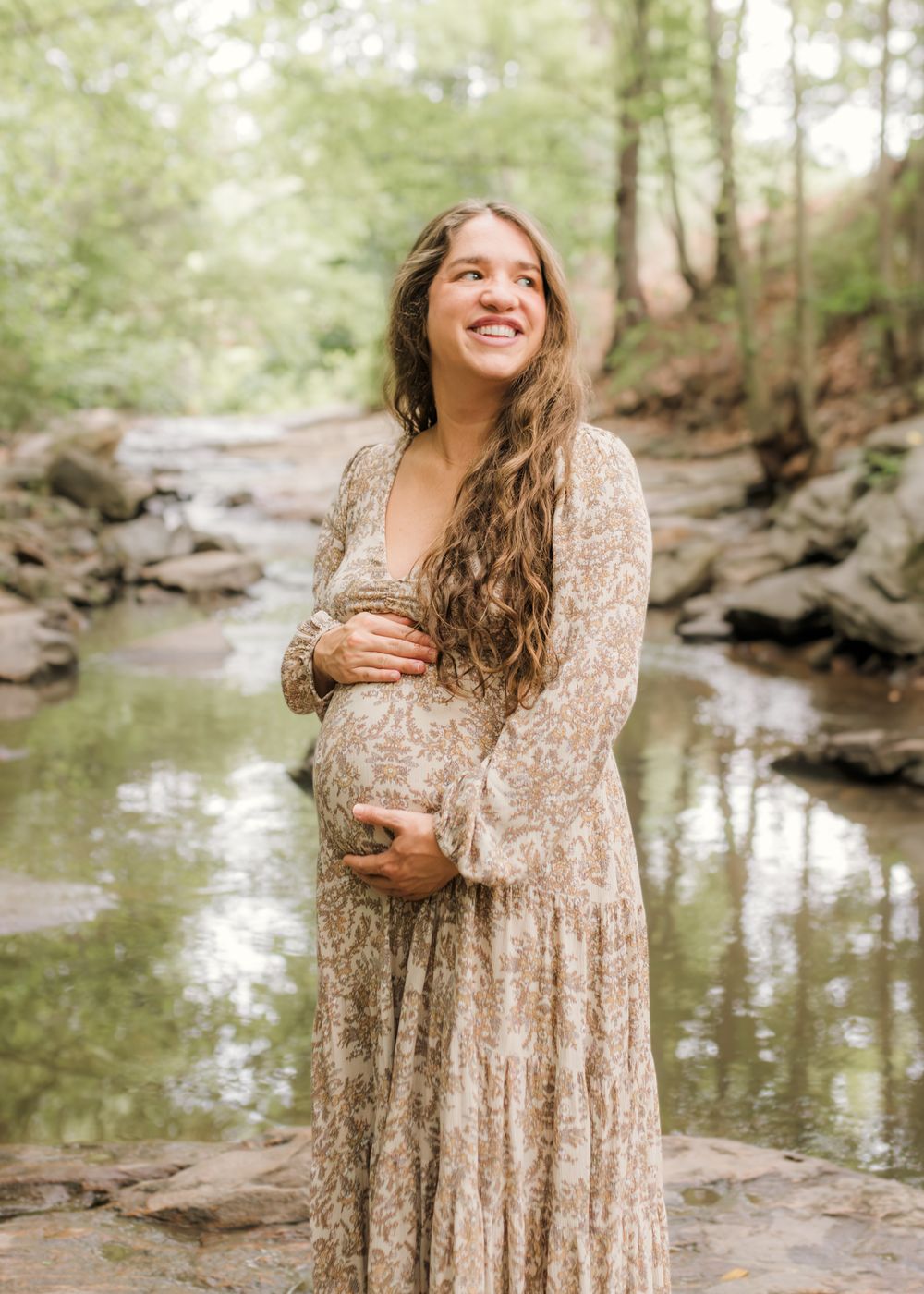 Smiling pregnant woman in floral maxi dress cradling baby bump by a serene forest creek.