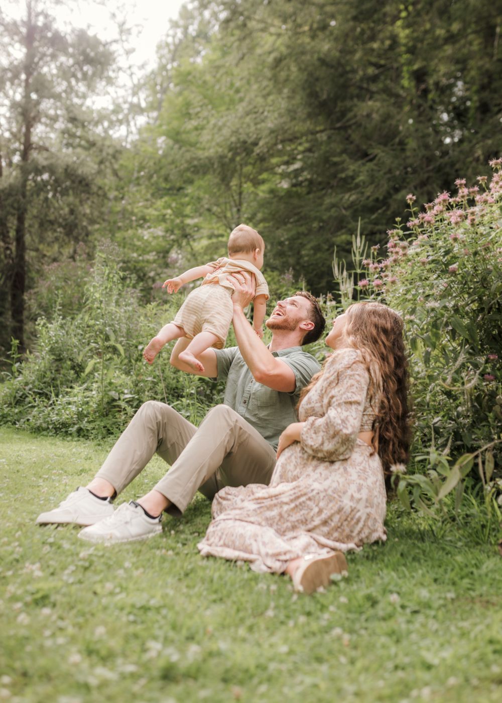 A joyful family sits on lush green grass, dad lifting baby in the air while mom watches, surrounded by blooming flowers.