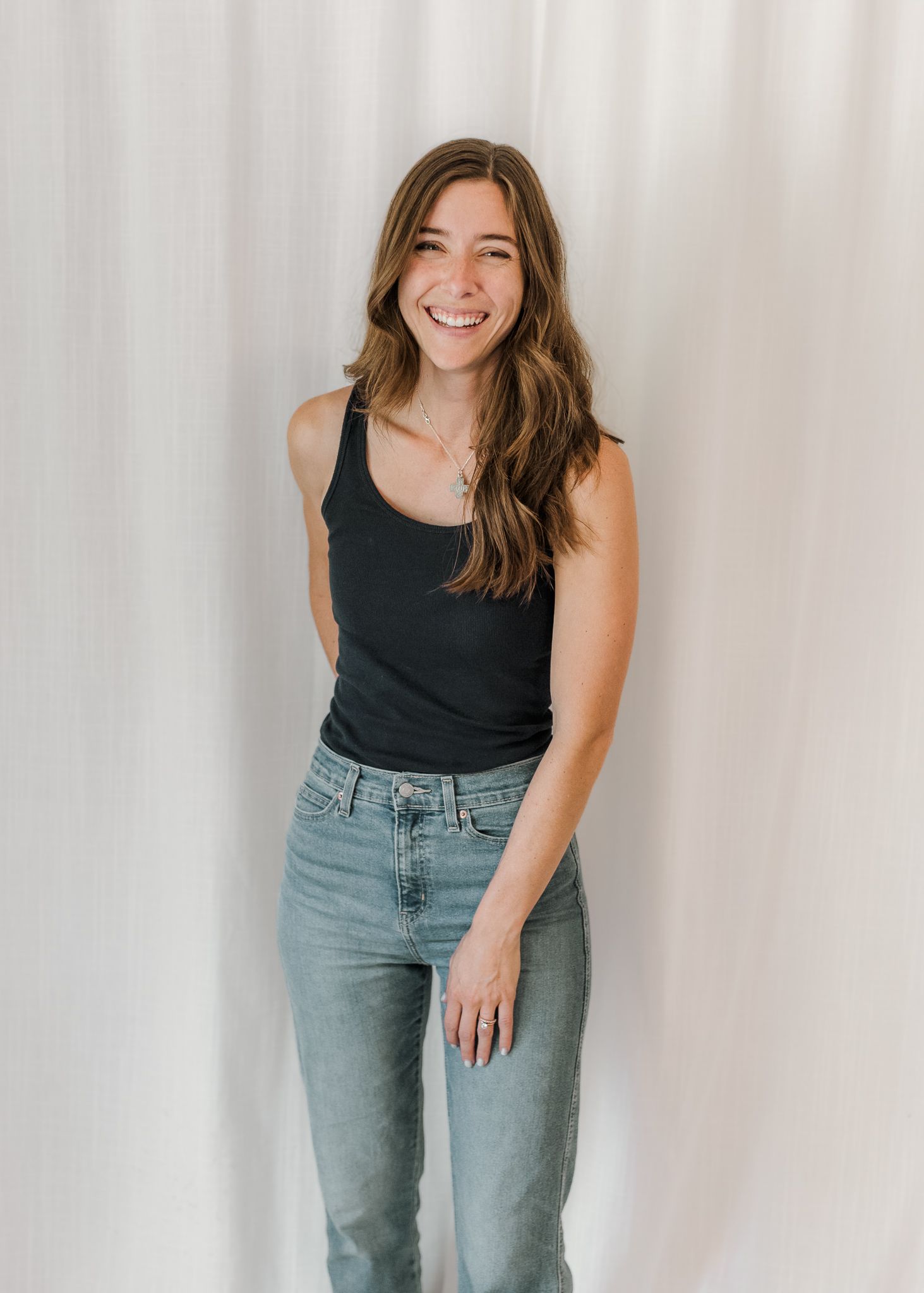 A person wearing a black sleeveless top and light blue jeans poses against a white backdrop in a casual photo series.