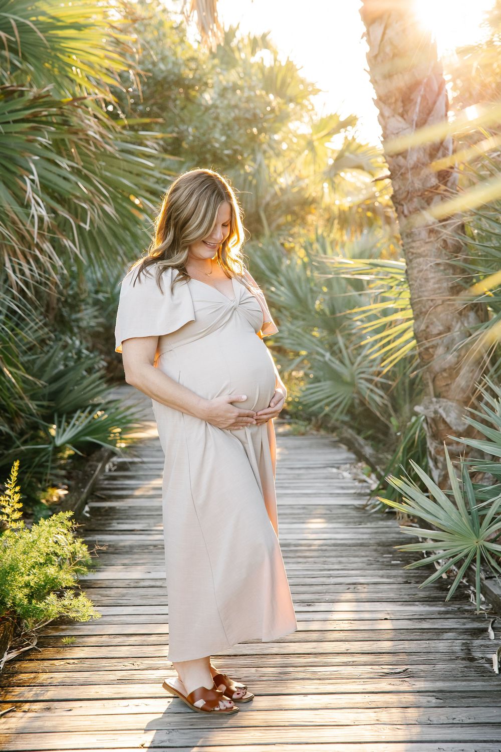 Pregnant woman in beige dress holding belly on wooden boardwalk surrounded by tropical palms in golden sunlight.