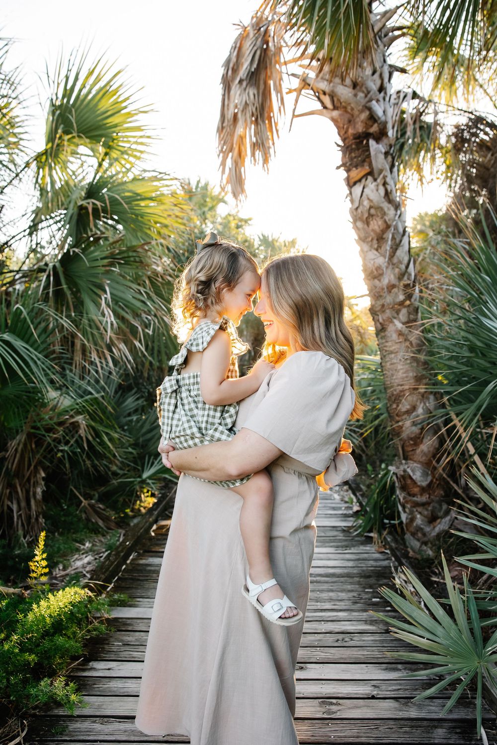 Mother holding young child on wooden boardwalk surrounded by palm trees in warm golden sunlight.