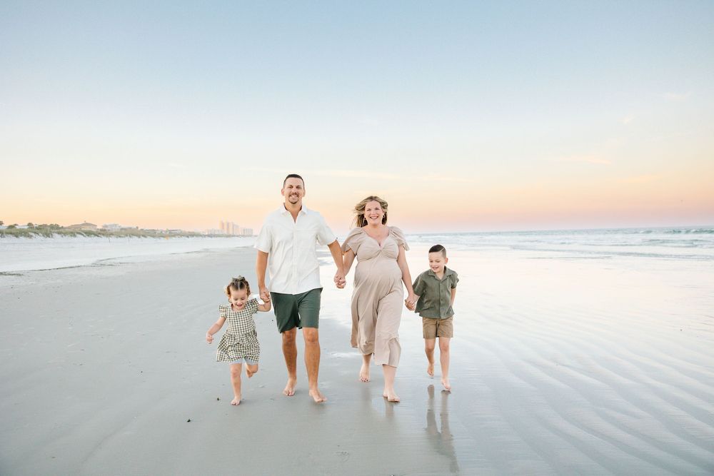 Family of four walks hand-in-hand on a sandy beach at sunset, with a pregnant mother in a flowing dress.