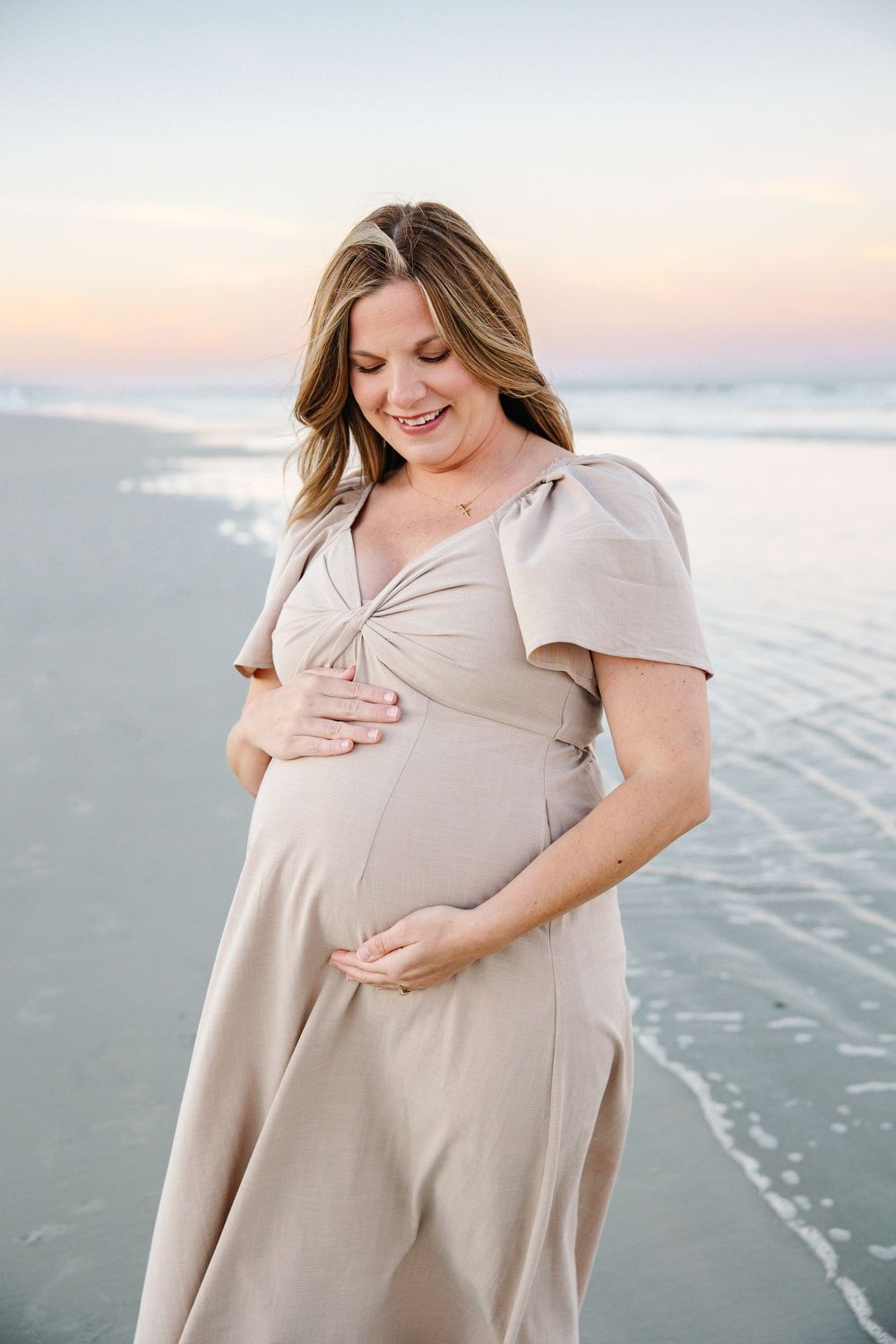 Smiling pregnant woman in beige dress cradling baby bump on beach at sunset.