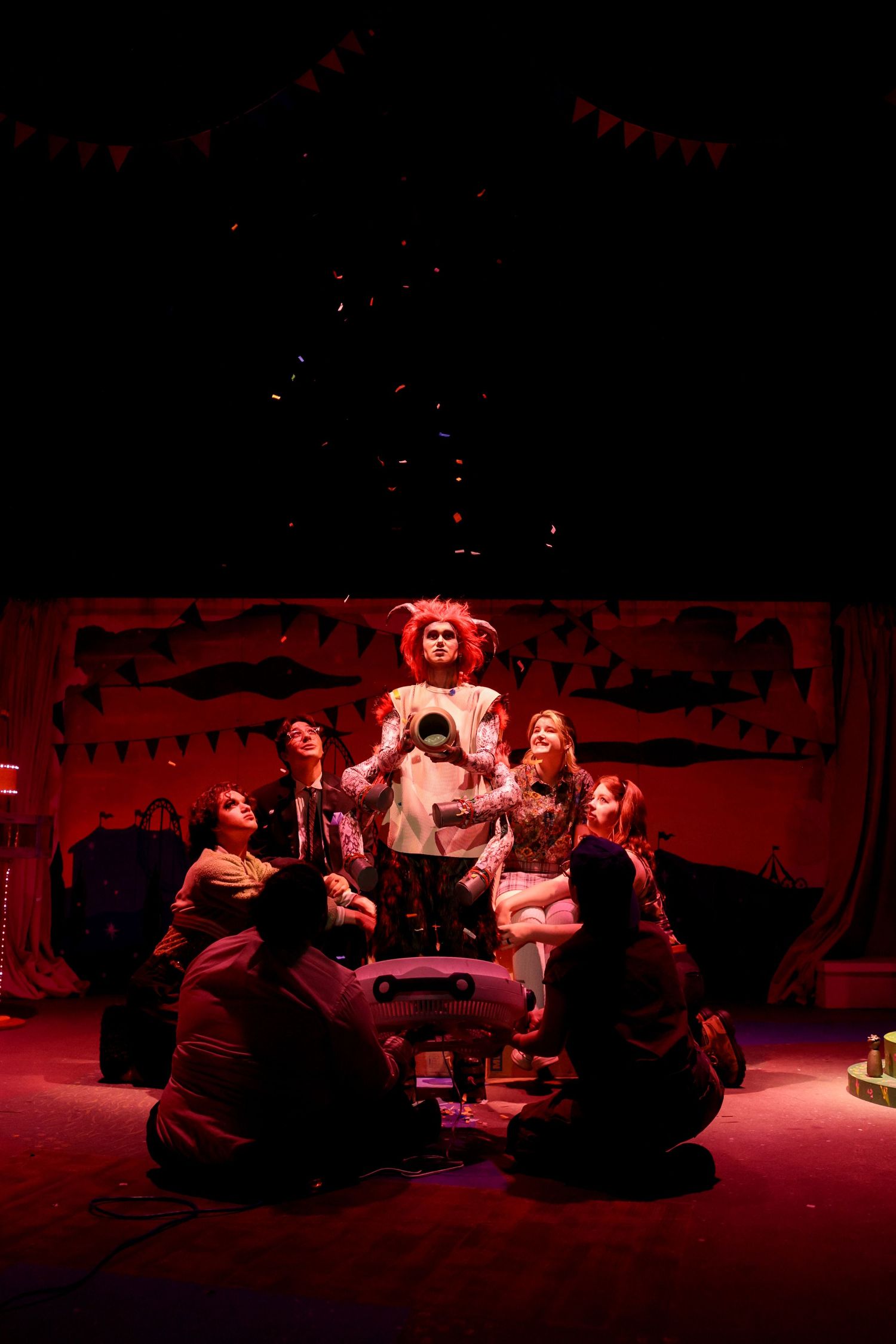 A theatrical performance bathed in red lighting shows performers gathered around in a circle on stage during a dramatic scene.