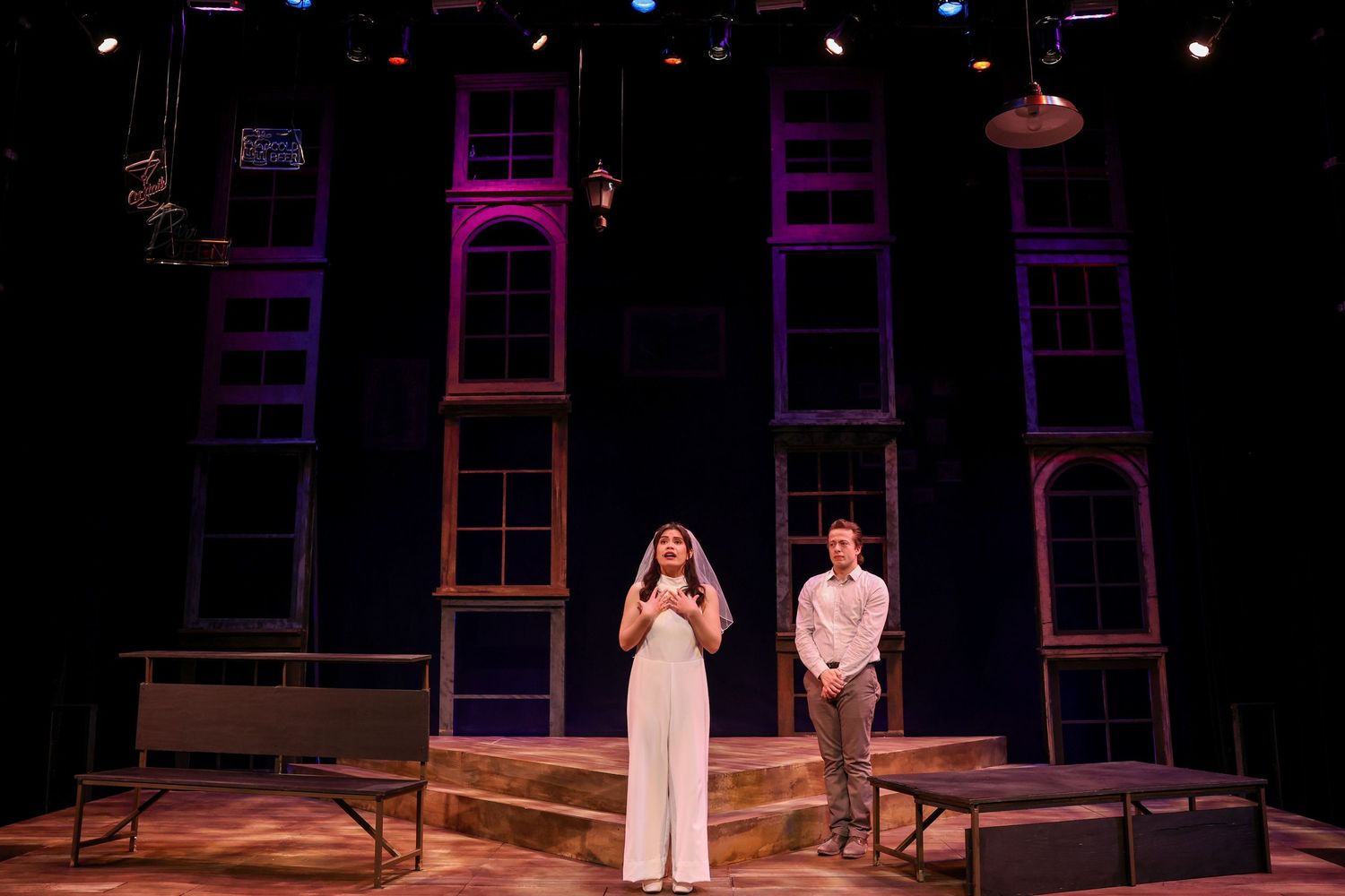 Performers stand on a theatrical stage with tall window-like set pieces illuminated in purple and orange lighting. Theatre photography.