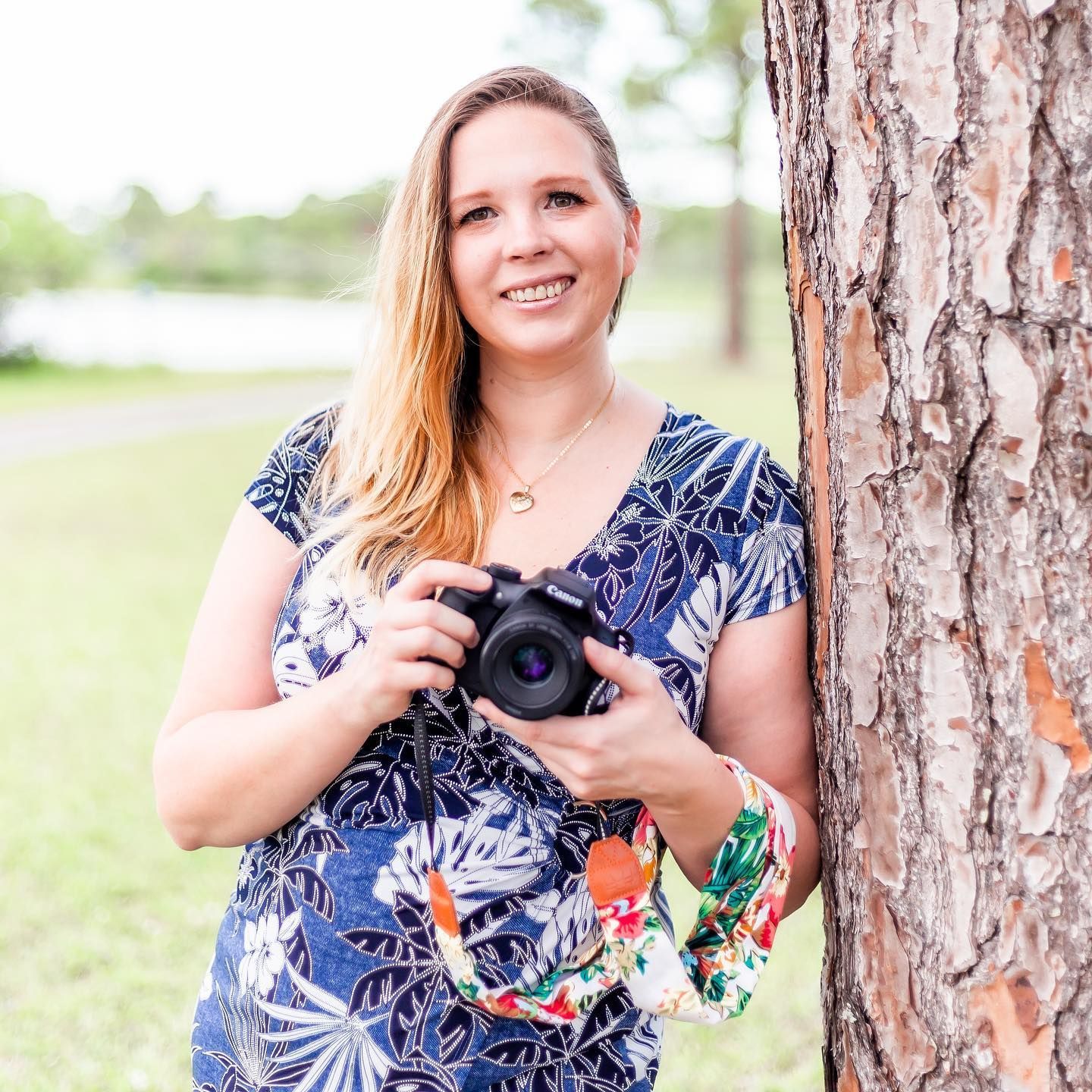 Photographer Kelsey from Photogenic Fox Photography in floral dress smiles while holding camera outdoors near pine tree.