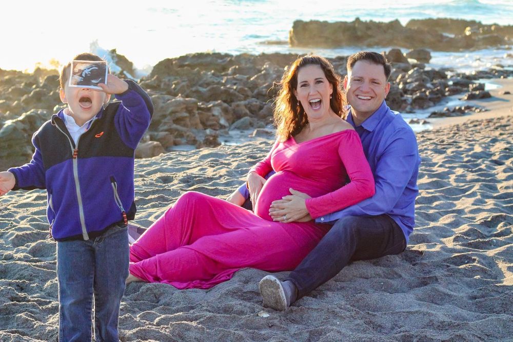 Pregnant woman in pink dress sits on beach with husband while excited young boy plays nearby at blowing rocks beach Jupiter fl.