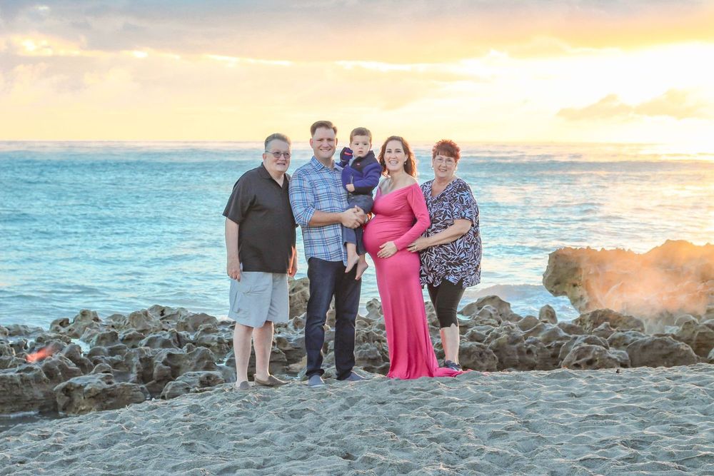 Family of five poses on rocky beach at sunset, pregnant woman in pink gown, ocean waves and colorful sky behind them at blowing rocks beach.