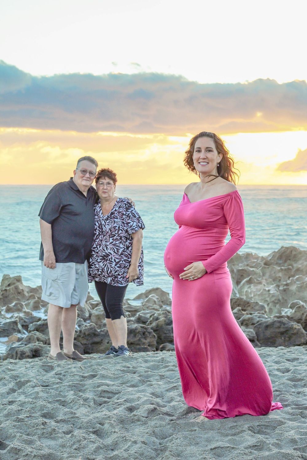 Pregnant woman in pink gown poses with couple on beach at sunset at blowing rocks beach Jupiter fl.