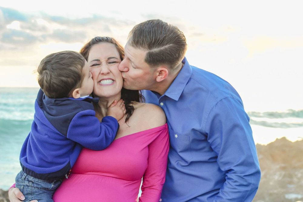 Pregnant woman in pink dress receives kisses from husband and young son on a beach at sunset at blowing rocks beach Jupiter fl.