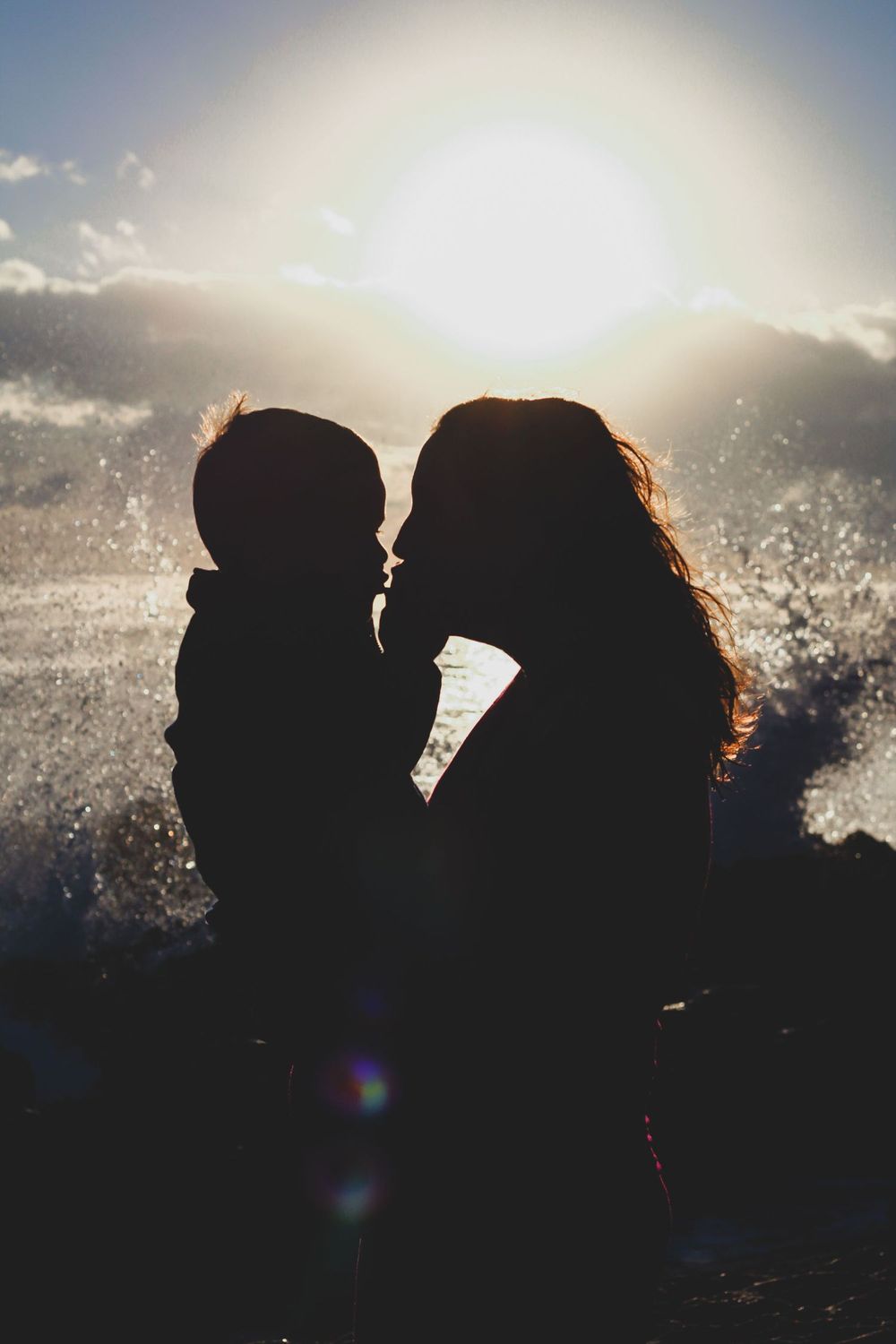 Silhouette of mother and child sharing a tender kiss against a stunning sunset with ocean waves splashing at blowing rocks beach Jupiter fl.