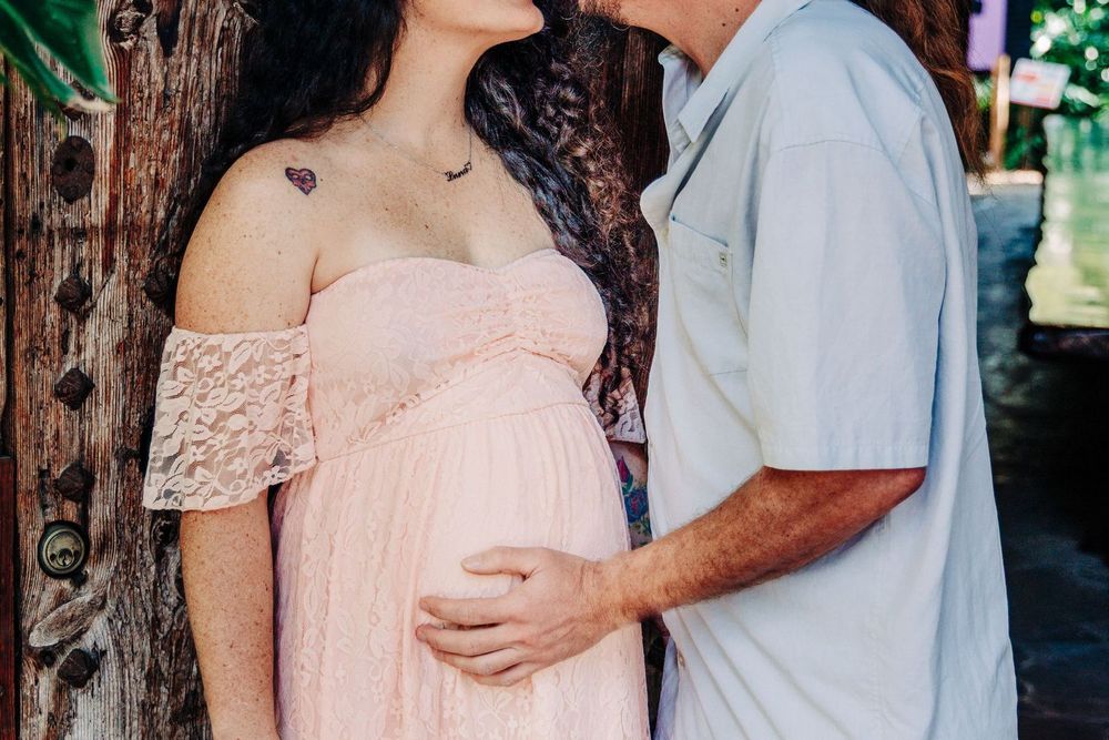 A pregnant woman in a pink lace off-shoulder gown poses with her partner outdoors during a maternity photo shoot at Mckee Botanical Gardens.