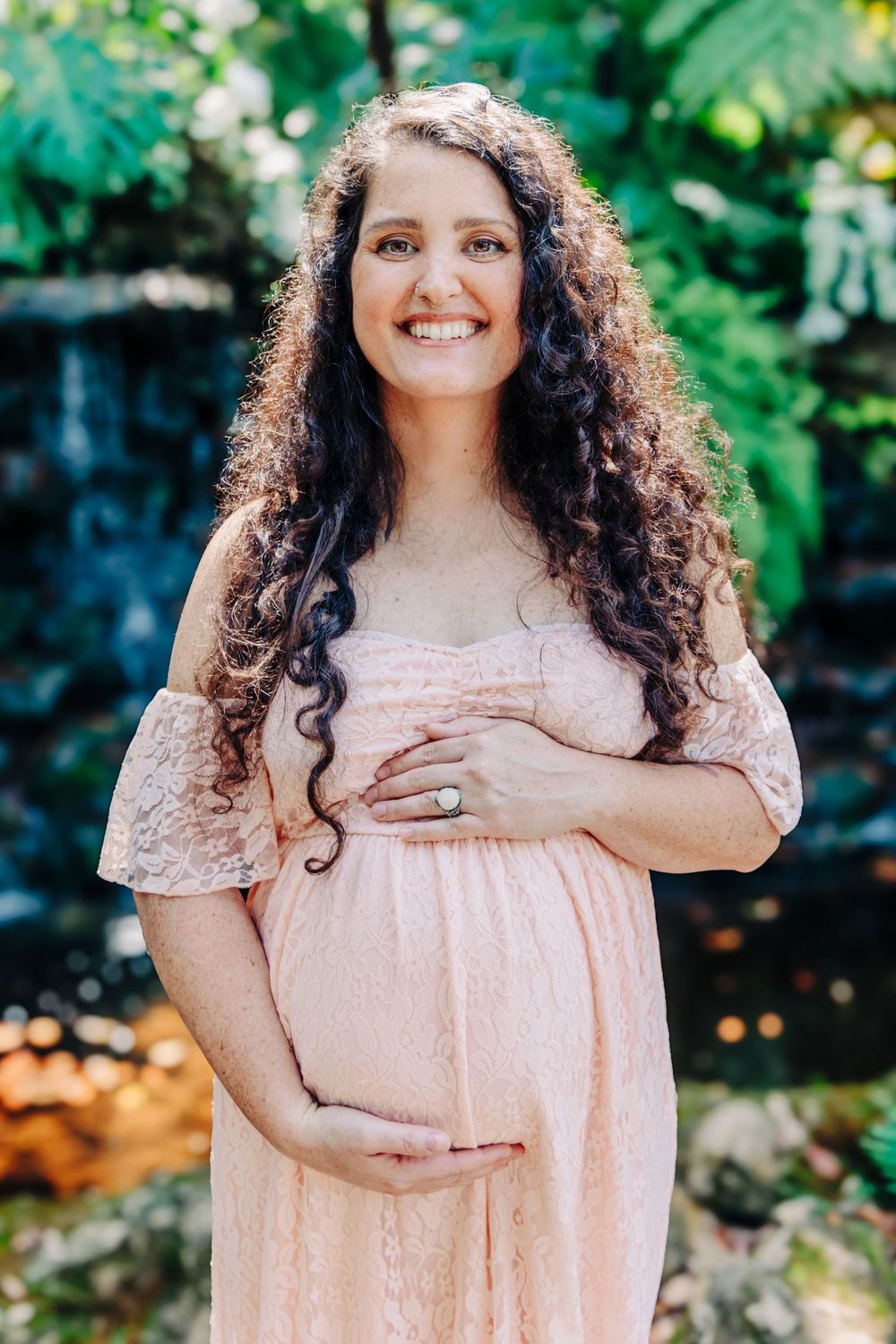 Smiling pregnant woman in pink lace off-shoulder dress cradling baby bump outdoors with lush greenery backdrop at Mckee Botanical Gardens.