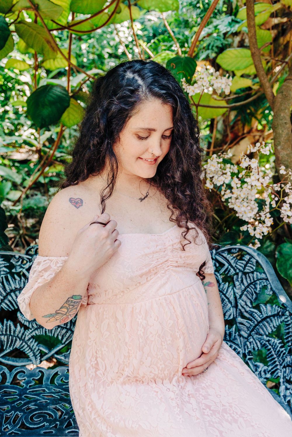 Pregnant woman in pink lace off-shoulder dress sits on ornate bench, cradling baby bump amid lush garden at Mckee Botanical Gardens.