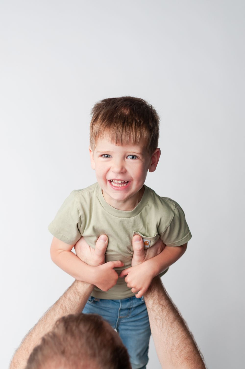A laughing toddler boy in a green shirt being lifted by an adult against a white background.