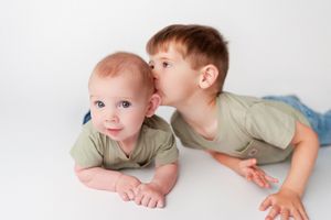 Two young brothers in matching green shirts, older boy kissing baby sibling on cheek on white backdrop.