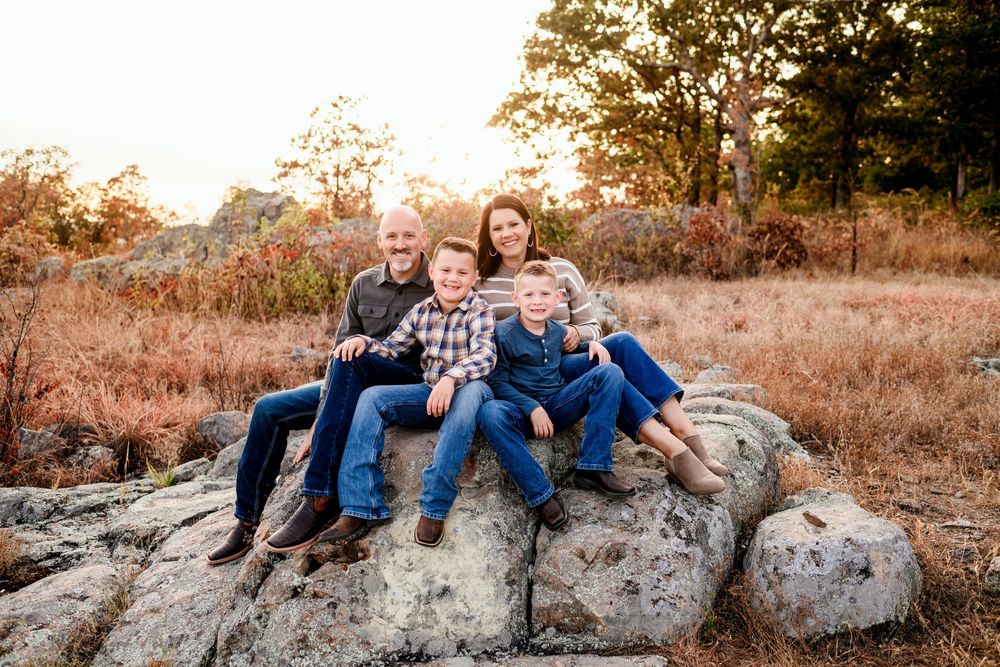 A family of four sits together on rocks in an autumn field during golden hour.
