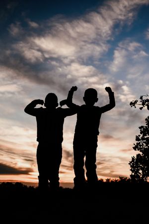 Silhouettes of two children flexing their muscles against a dramatic sunset sky.