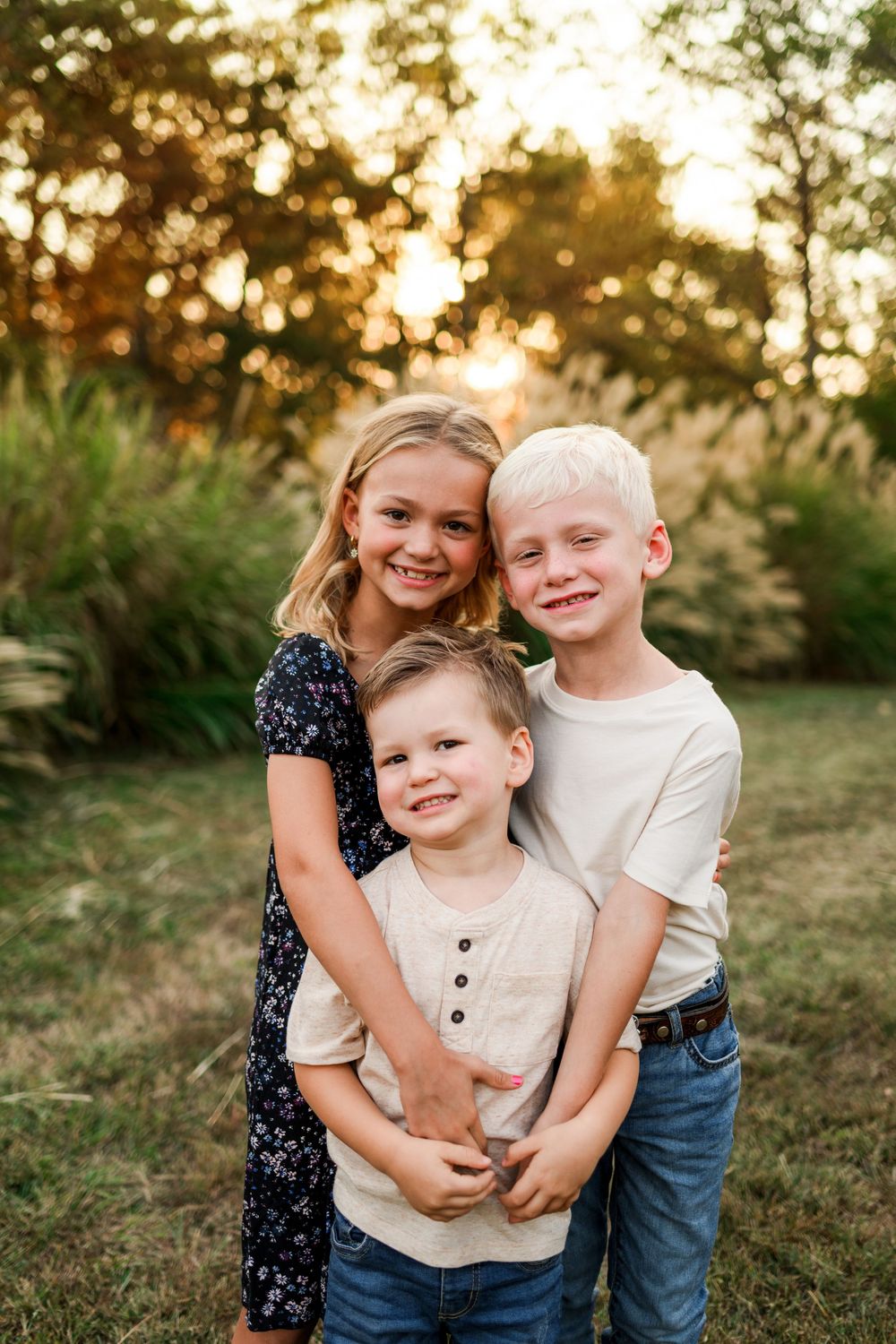 Three smiling siblings hugging together outdoors in golden sunset light among green trees.