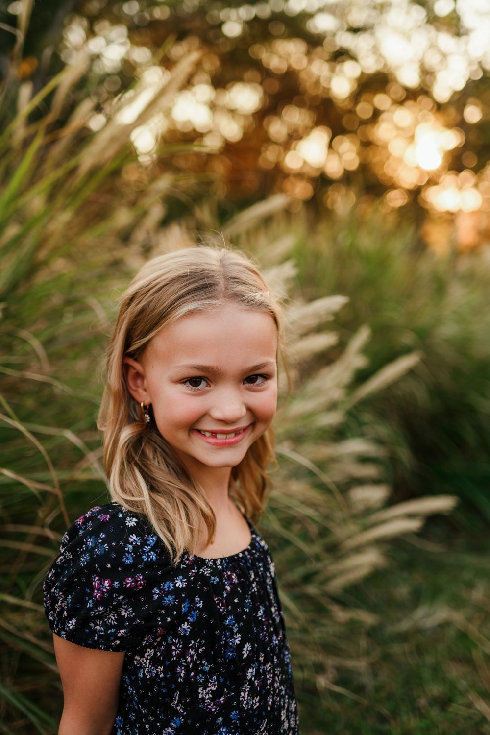 Smiling young blonde girl in floral dress posing outdoors in warm golden sunset light.