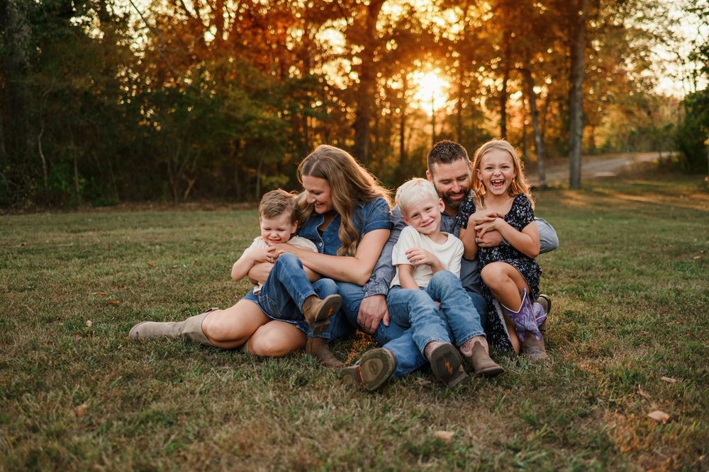 Happy family of five laughing and sitting together on grass during golden hour sunset.