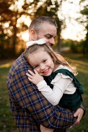 A father tenderly embraces his smiling young daughter in a green dress at sunset outdoors.