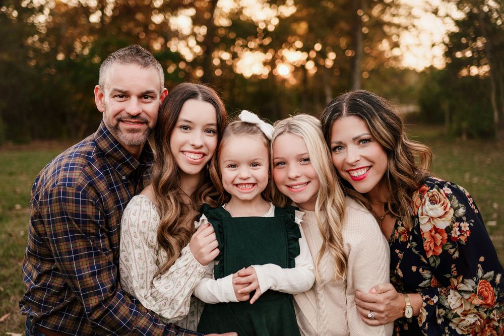 A family of five smiles together outdoors at golden hour, with three girls and two parents.