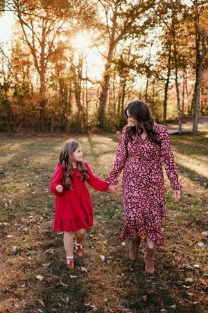 Mother and daughter in red floral dresses holding hands walking in autumn sunlit forest.
