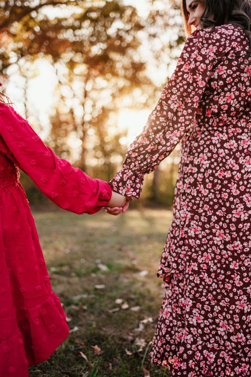 Mother and young daughter holding hands in red and floral dresses in autumn park.