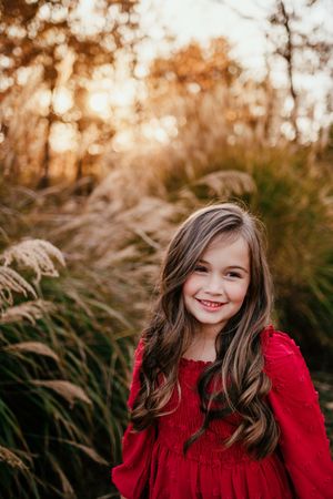 A smiling young girl with long brown hair wearing a red dress poses outdoors at golden hour.